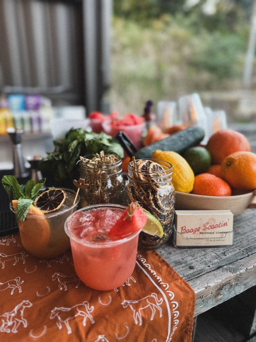 A pink strawberry cocktail on a printed runner with fresh citrus, cucumbers, and mint behind.