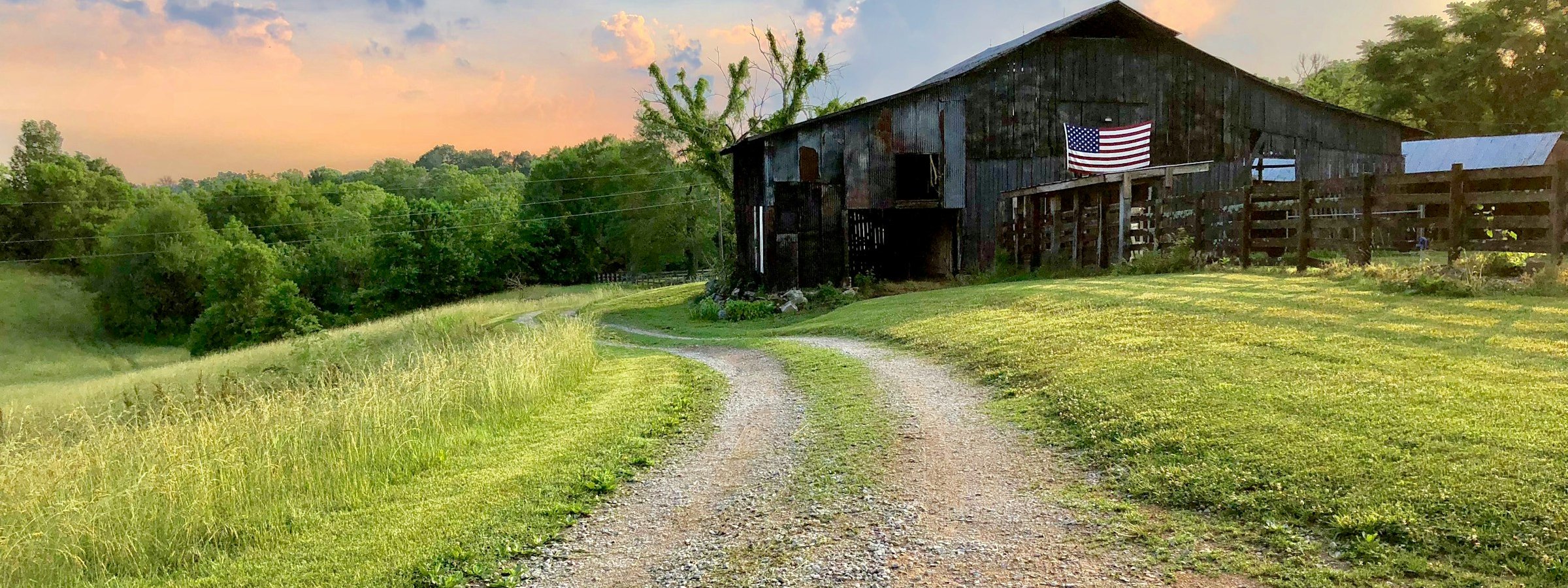 A gravel farm road curves toward a weathered wooden barn flying a US flag at sunset outside Columbia.