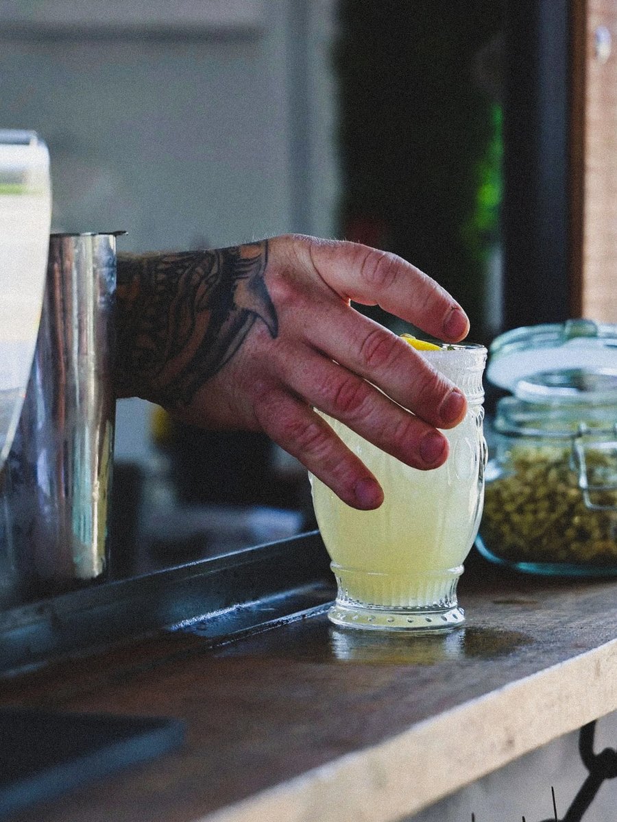 A tattooed hand reaches through the trailer window for a light cocktail at golden hour, purple flowers hanging alongside.