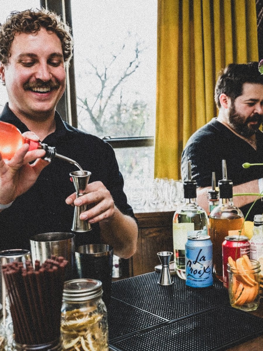 A bartender in black pours a red cocktail into a jigger, another arranges flowers at the wooden bar behind him.