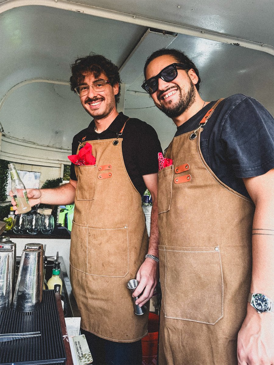 Two Booze Scootin' bartenders in tan aprons and black shirts smile behind the bar setup inside the trailer.