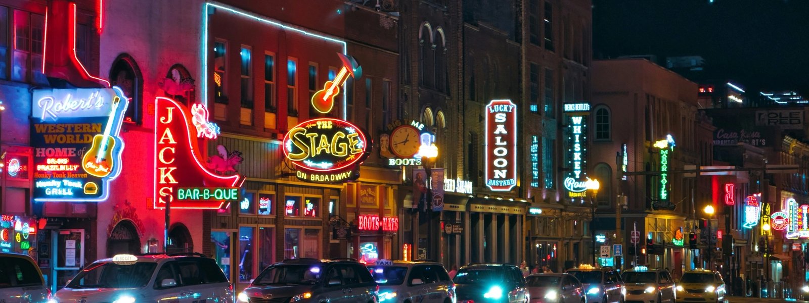 Lower Broadway at night, honky-tonk neon along the block: Robert's Western World, Jack's Bar-B-Que, The Stage on Broadway, Lucky Bastard Saloon, Whiskey Row. Taxis lined up along the curb.