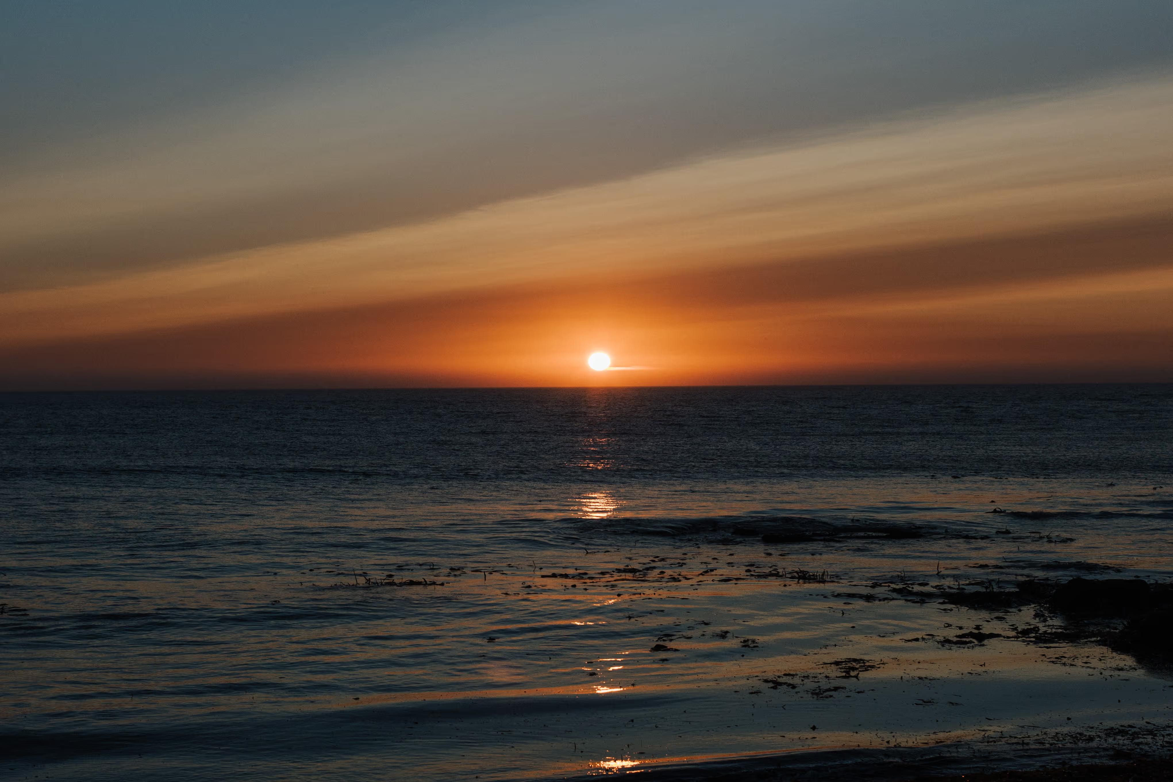 Sol a pôr no horizonte sobre um mar calmo com reflexos dourados na água e céu com nuvens coloridas.