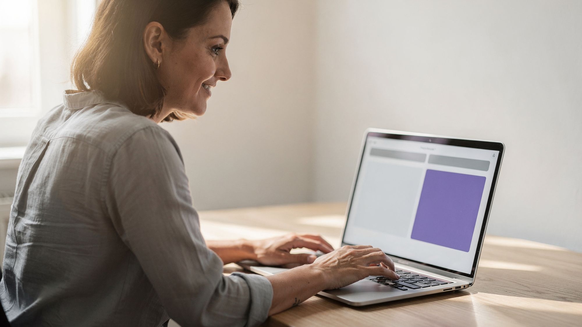 A woman types on a laptop at a light wood desk, the screen showing a spacious layout of soft colour blocks with one block in violet.