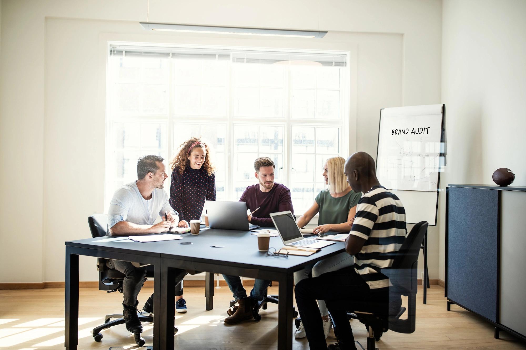 Dancing Pels team collaborates with a client around a table during a brand audit workshop, with a whiteboard displaying 'BRAND AUDIT' visible in the background.