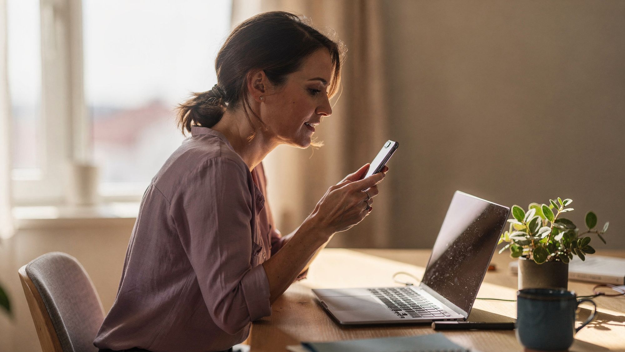 A woman sits at a light wood desk with an open laptop in front of her and holds her phone up in one hand, looking at the phone screen.