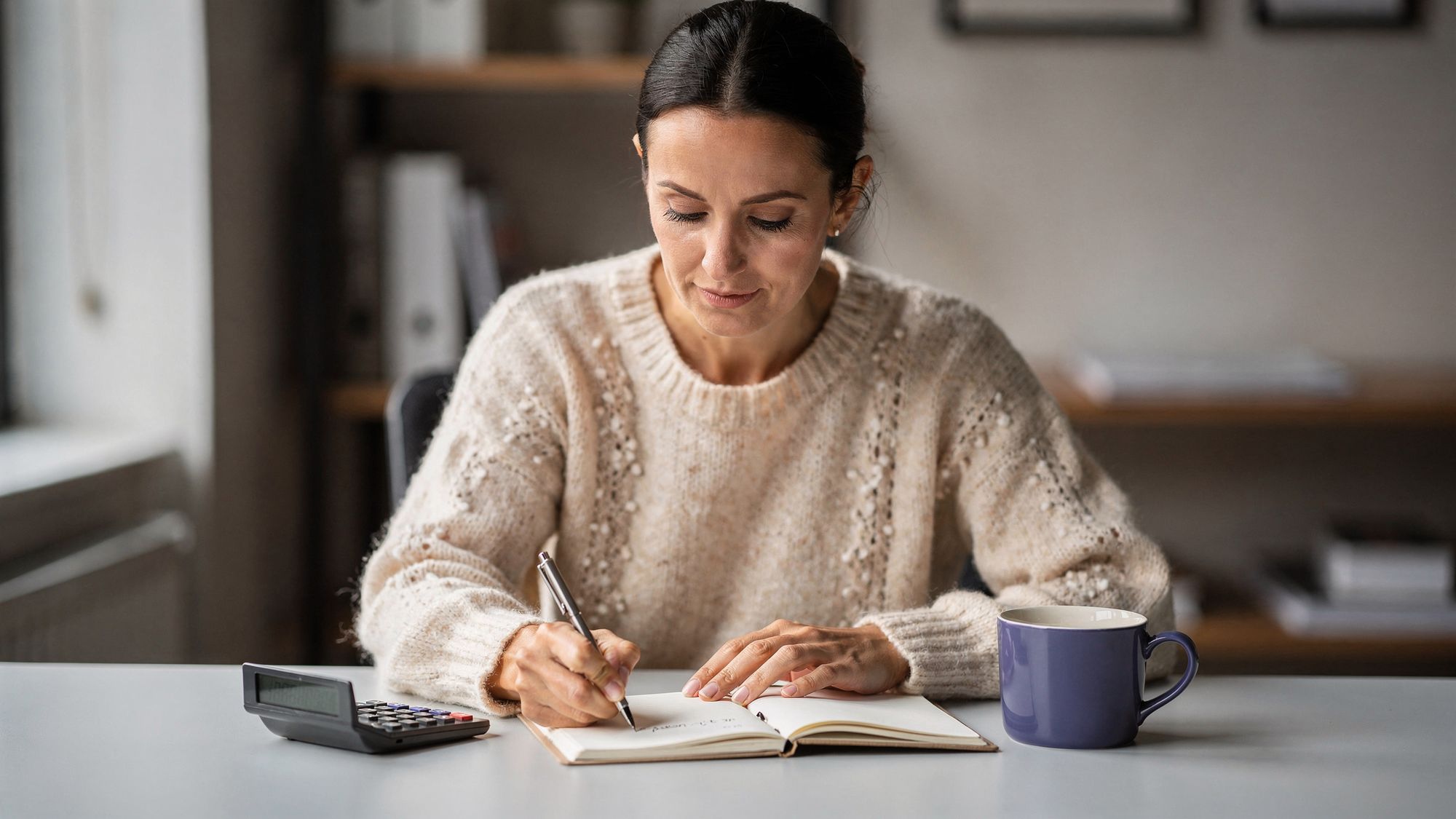 A woman in a cream knit sweater writes a short line in a notebook at a table, with a small calculator under her left hand and a violet mug beside her, in morning light.