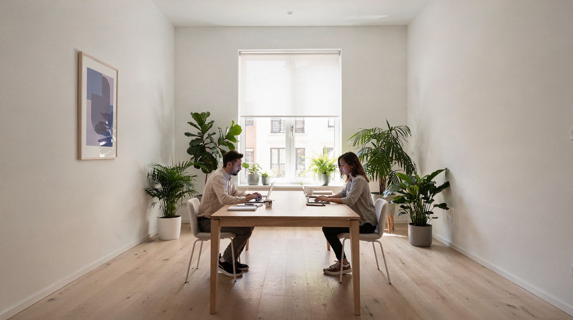 Two people work on laptops at opposite ends of a long wooden table in a bright studio office full of plants, lit by a large window.