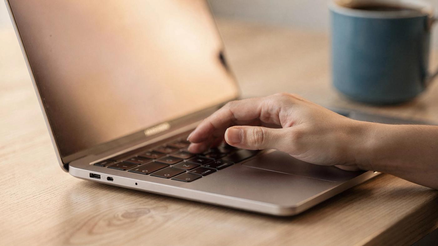 A close view of a hand resting on a laptop trackpad with the index finger lifted, lit by the warm glow of the screen.