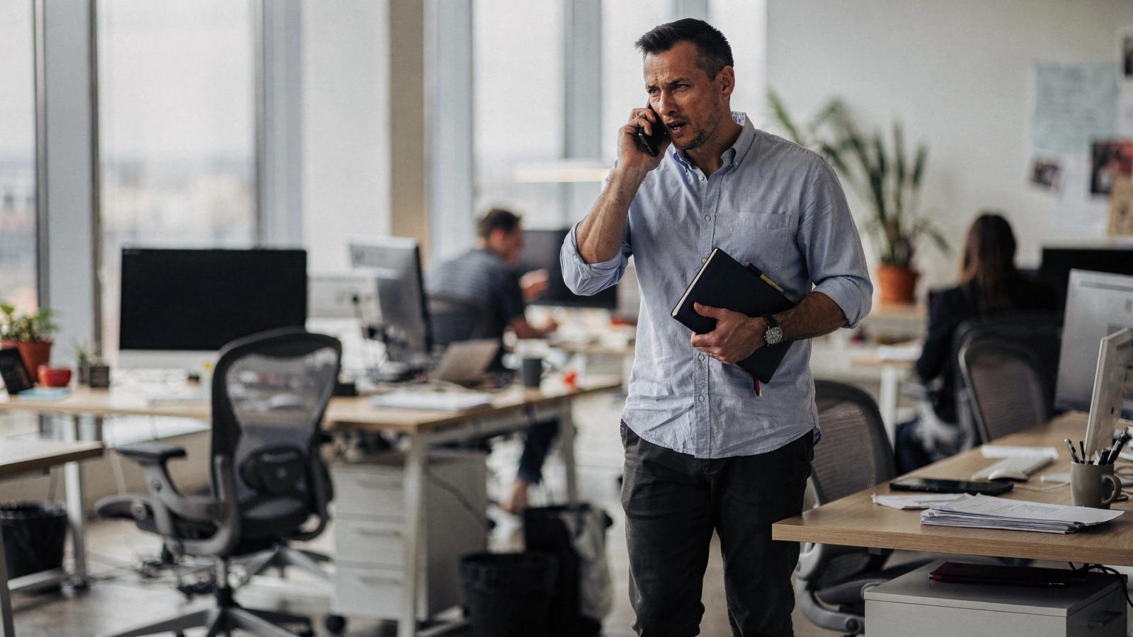 A man in a light blue shirt walks through an open plan office with a phone held to his ear and a dark notebook under his arm, colleagues working at desks behind him.