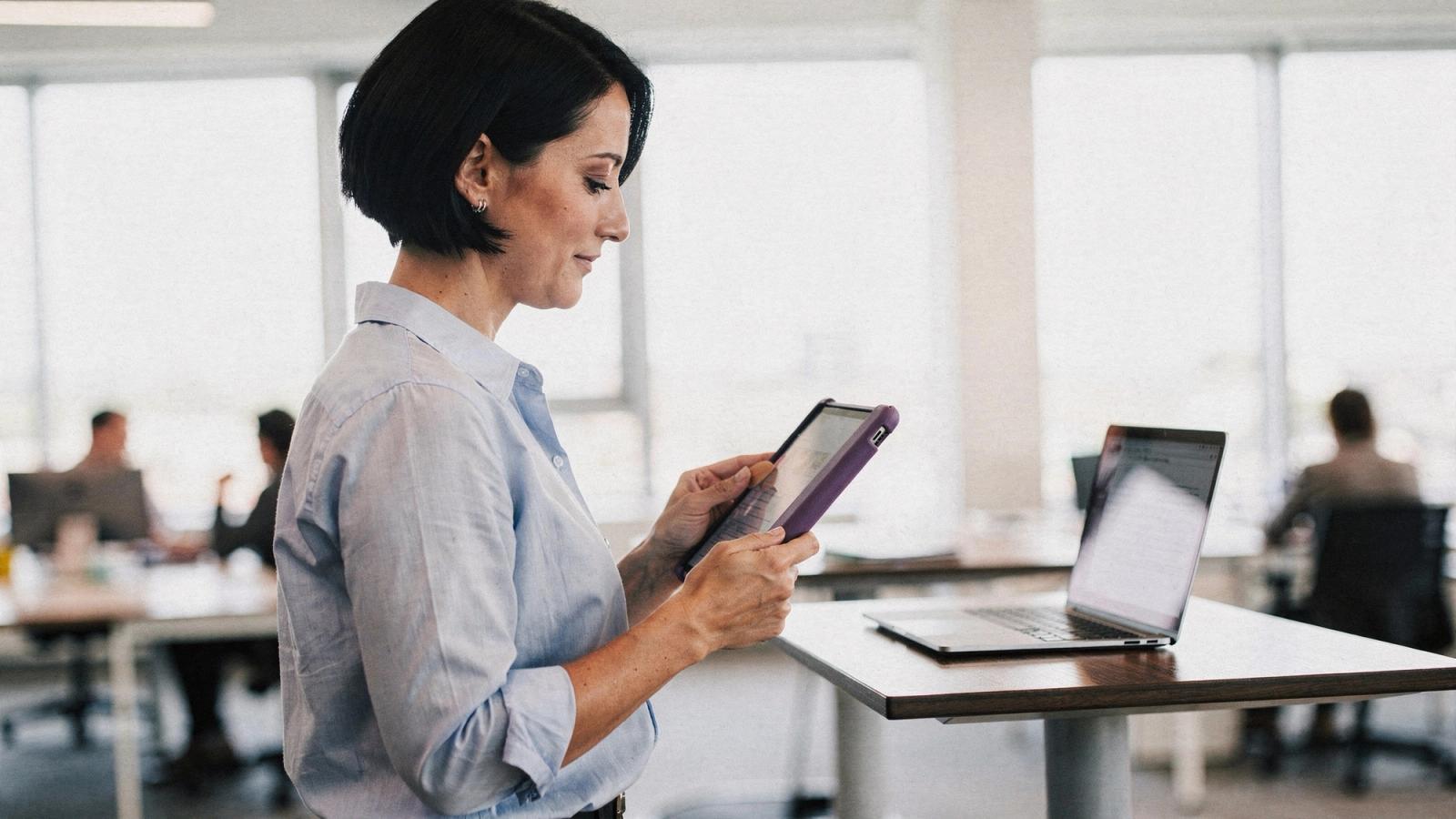 Una mujer de pelo corto negro y camisa azul clara, de pie ante un escritorio alto en una oficina luminosa, toca una tableta con funda violeta junto a una laptop abierta.