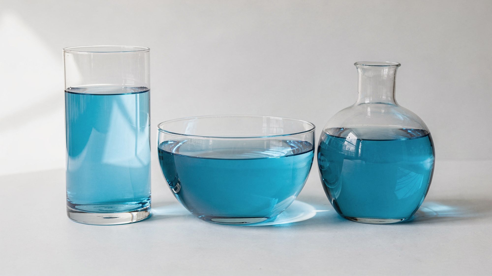 Un cilindro alto, un tazón ancho y un matraz redondeado están en fila sobre una superficie lisa, cada uno con la misma agua azul que toma la forma de su contenedor, bajo una luz suave de día.