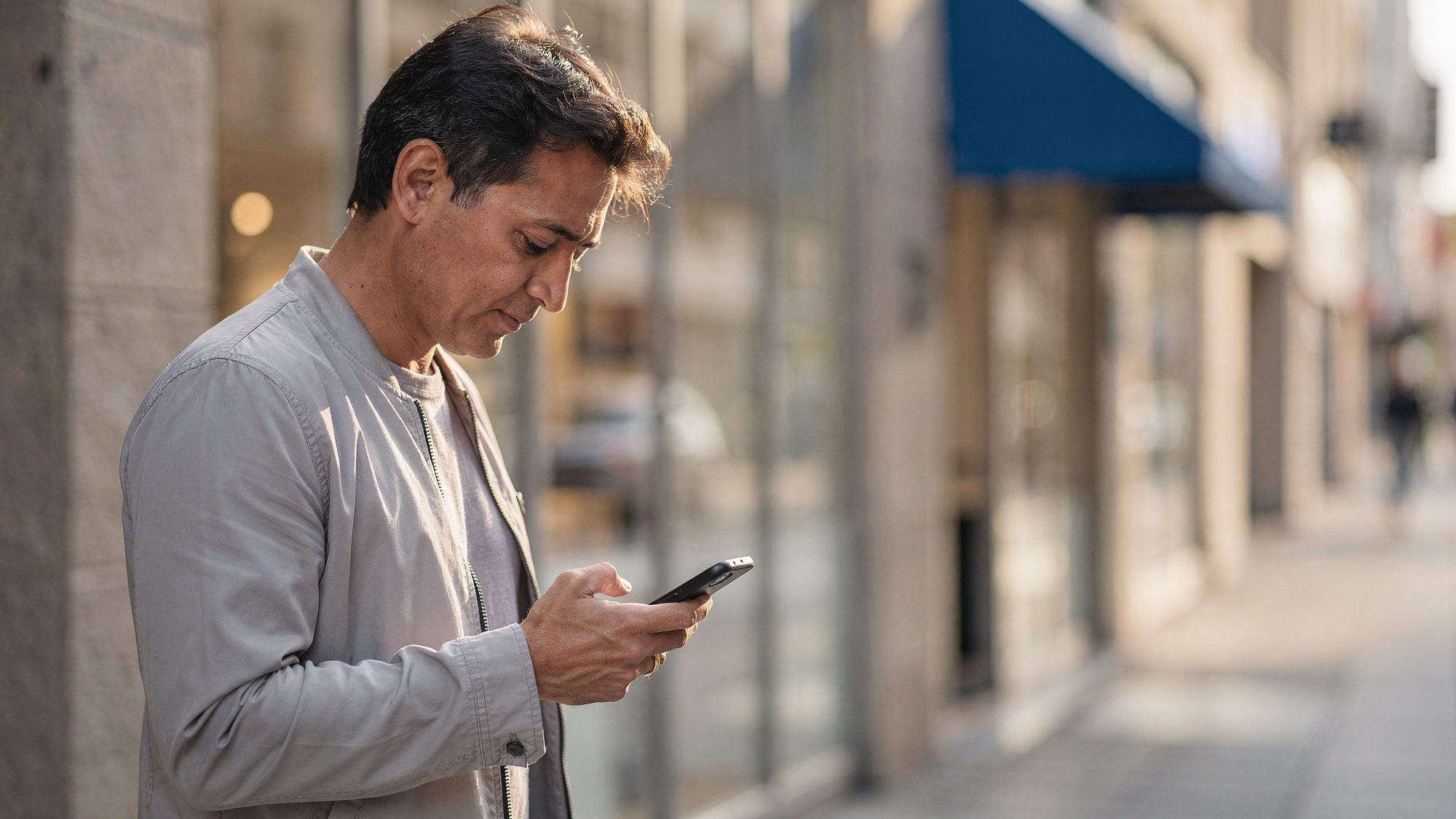 A man stands on a sunlit city sidewalk looking at his phone, with a modern glass storefront out of focus behind him.