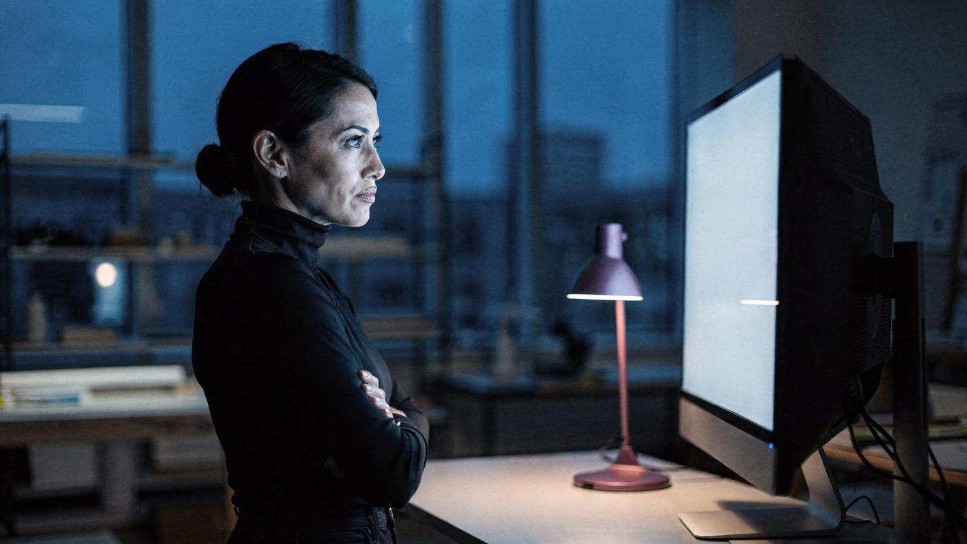 A woman in a black turtleneck stands with her arms crossed in a studio at dusk, lit by the glow of a large screen that faces away from the camera, with a small desk lamp beside her.
