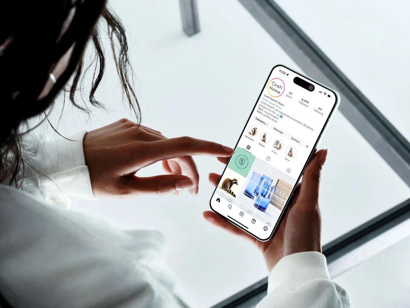 Person interacting with a mobile app interface displaying a curated home goods brand with product grid, navigation, and social features.