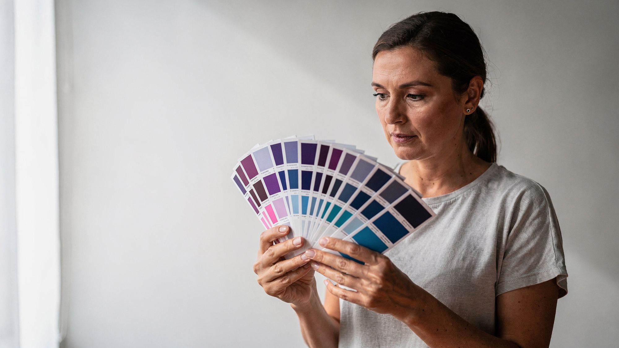 A woman holds a fan of printed colour swatch cards open in front of a white wall, running through violets and blues, and studies them.