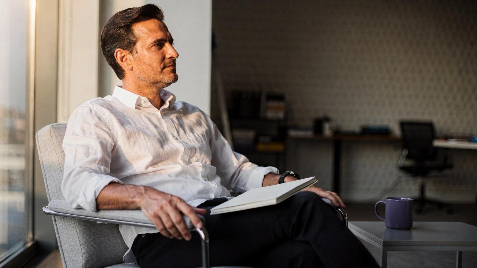 A man in a white linen shirt sits in a low chair beside a window with an open notebook resting on his lap, looking out into the late afternoon light, a violet mug on the side table next to him.
