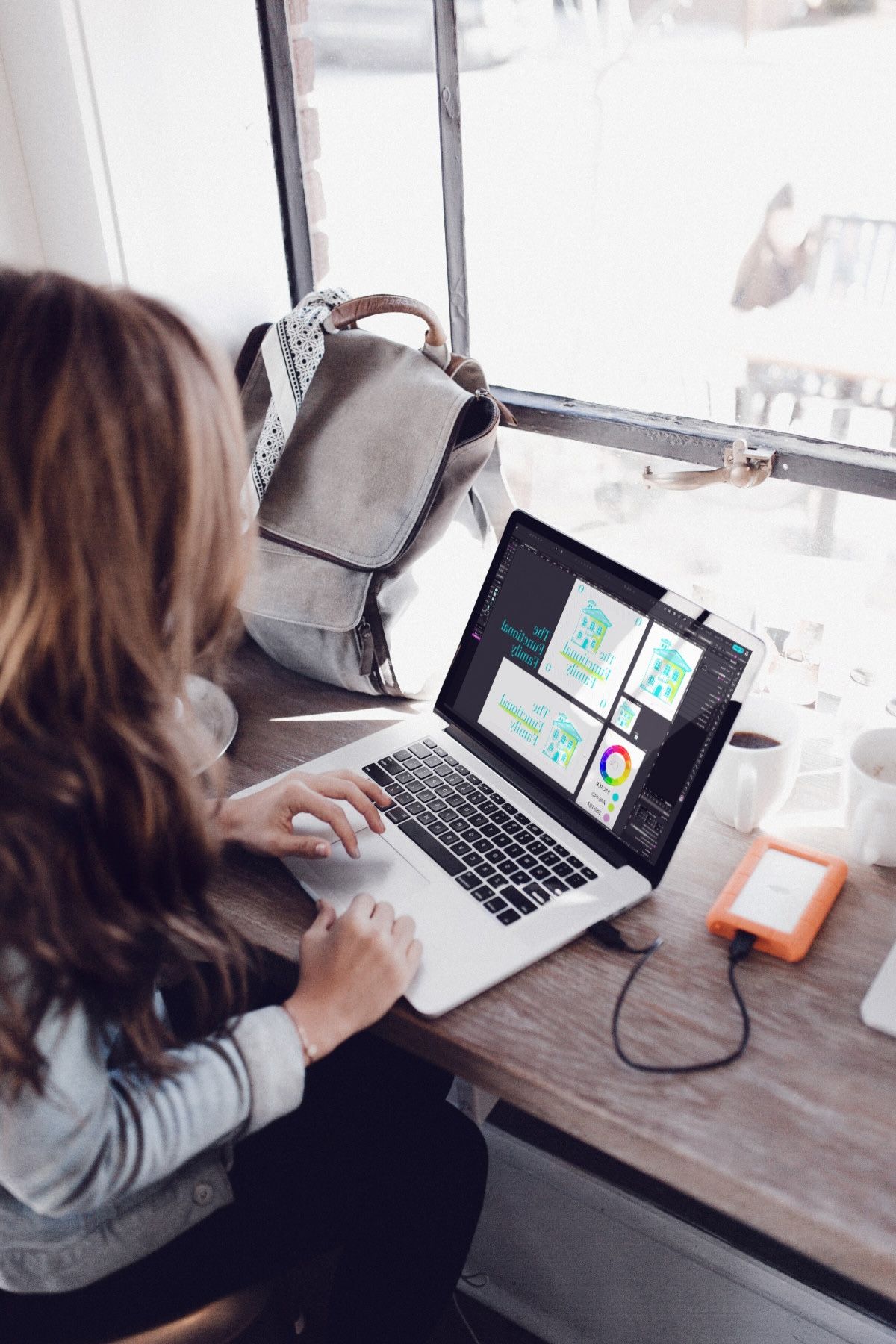 Designer working on a new brand on laptop by a bright window, with design assets and notebooks nearby.