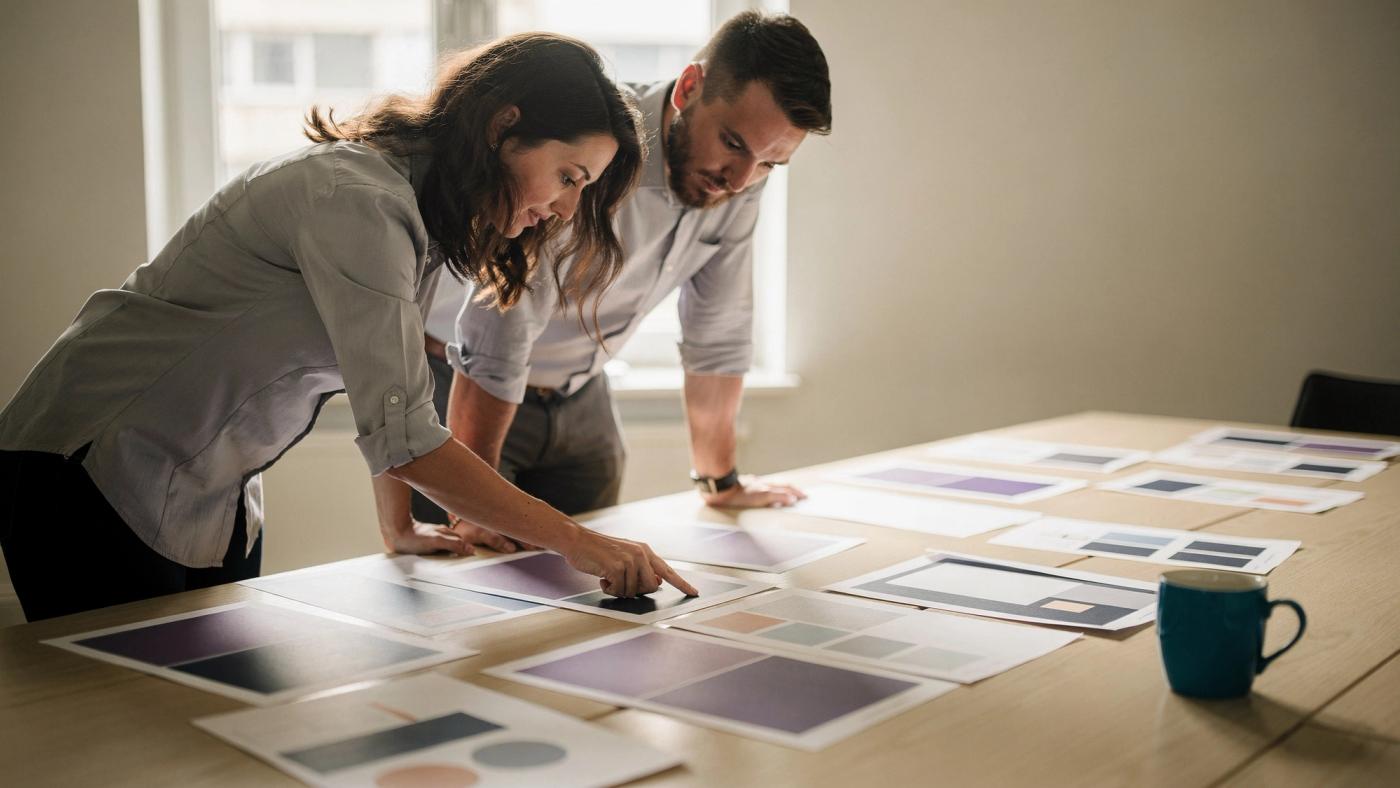 Two colleagues lean over a long table covered with printed sheets of colour blocks and shapes, one of them pointing at a single sheet.