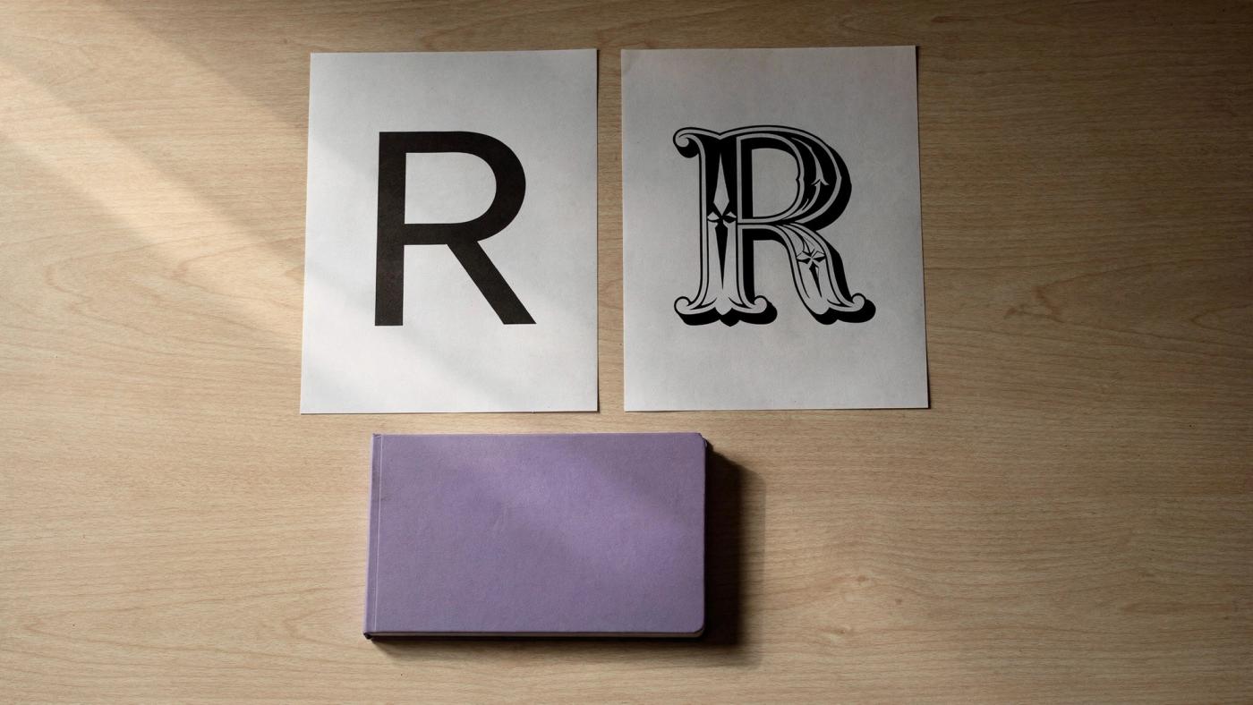 Two printed type specimen sheets lie side by side on a light wood table: the letter R set in a clean geometric sans serif, and the same letter set in an ornate Victorian serif.