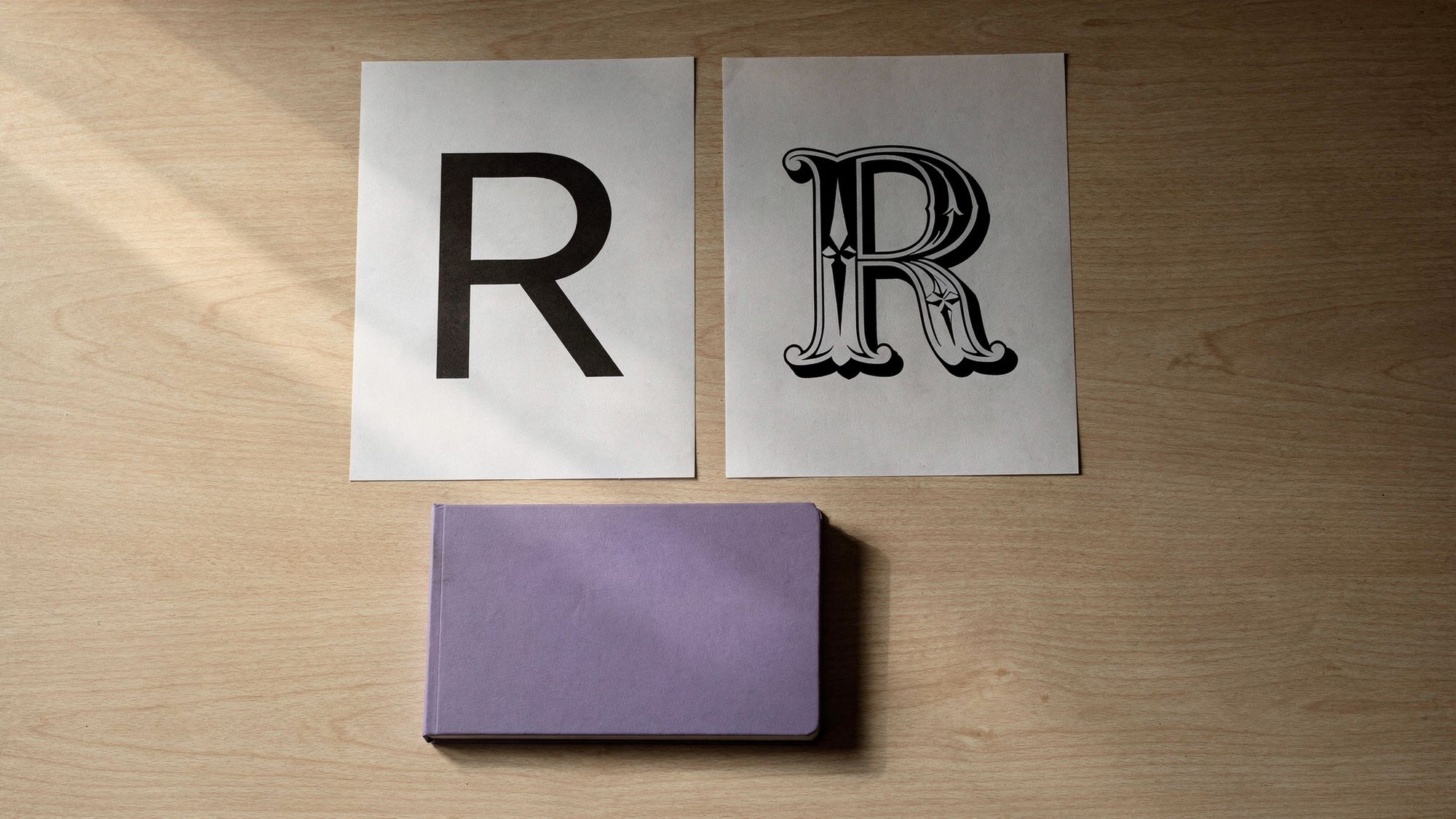 Two printed type specimen sheets lie side by side on a light wood table: the letter R set in a clean geometric sans serif, and the same letter set in an ornate Victorian serif.
