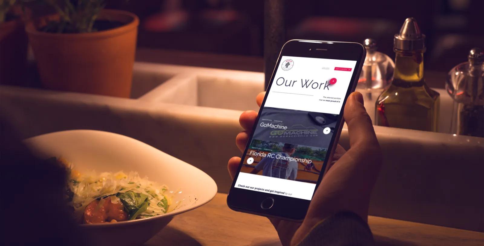 A person holds a smartphone displaying a Dancing Pels portfolio website with project examples, seated at a dining table with food and tableware nearby.