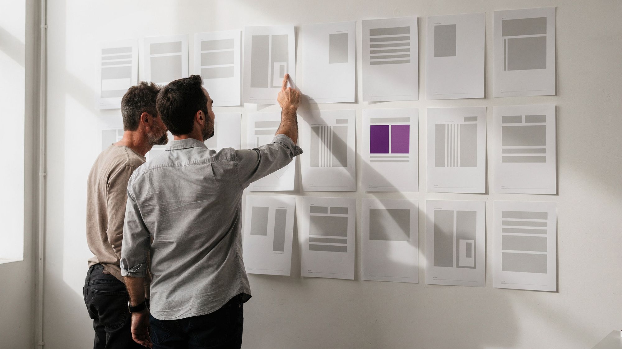 Two designers stand facing a white wall covered with a grid of printed page layouts, one of them reaching up to point at a single sheet.