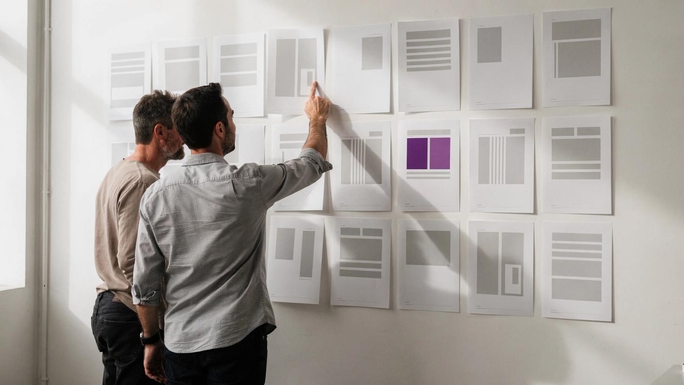 Two designers stand facing a white wall covered with a grid of printed page layouts, one of them reaching up to point at a single sheet.