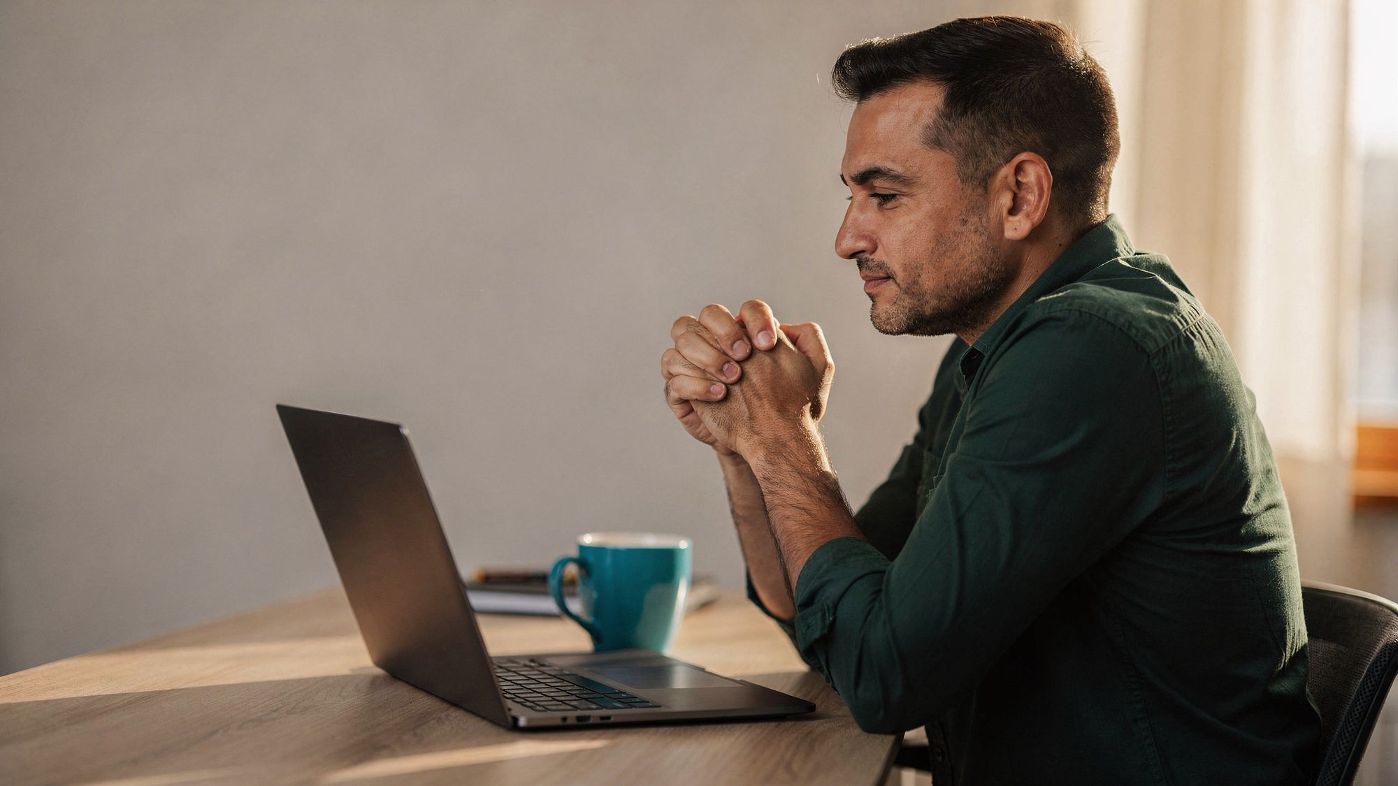A man in a dark green shirt sits at a light wood desk with his fingers laced together, looking at a laptop screen that faces away from the camera.