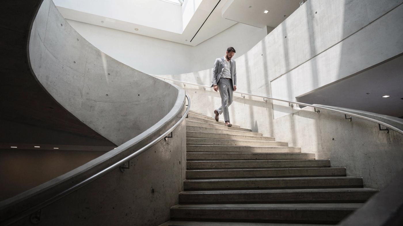 A man in a light grey suit walks down a sculptural curved concrete staircase inside a bright modern building, lit by a skylight above.