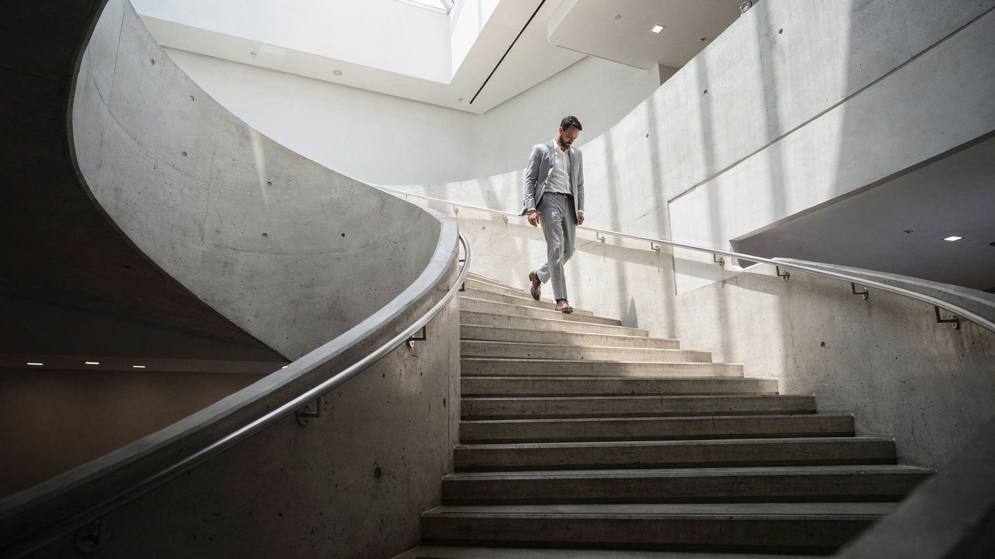 Un hombre con traje gris claro baja por una escalera de concreto curva y escultural dentro de un edificio moderno y luminoso, iluminado por un tragaluz desde arriba.