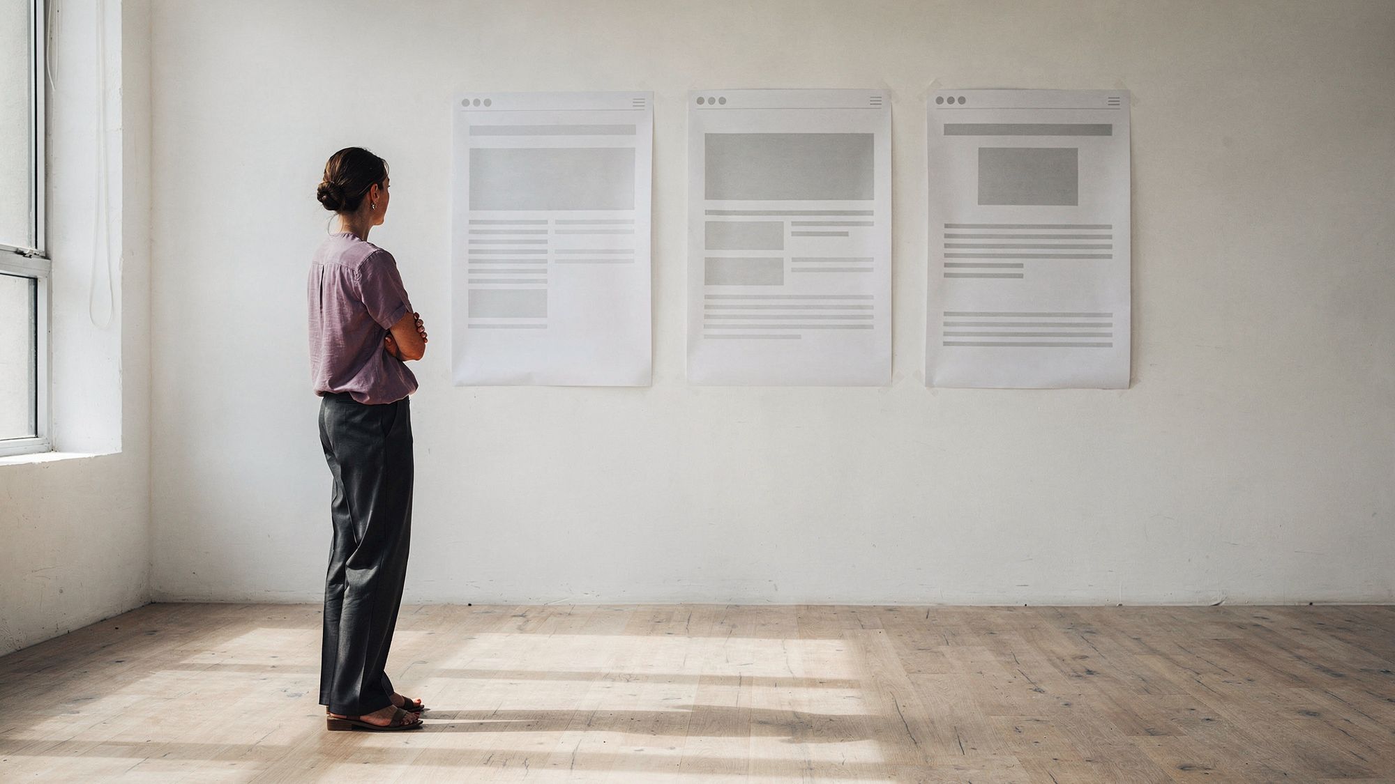 A woman stands back from a white studio wall where three printed website wireframes are taped in a row, arms folded, studying the middle one.