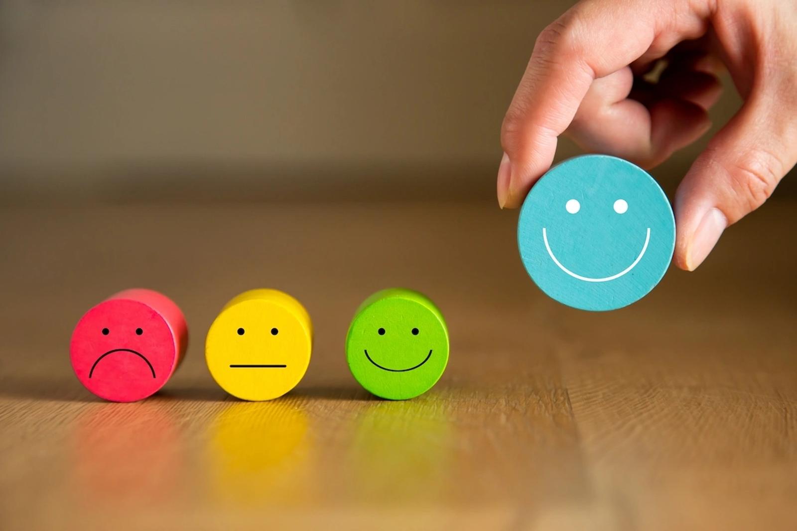 A hand holds a turquoise smiley face above a row of colorful emotion faces, representing how Dancing Pels helps clients achieve positive outcomes.