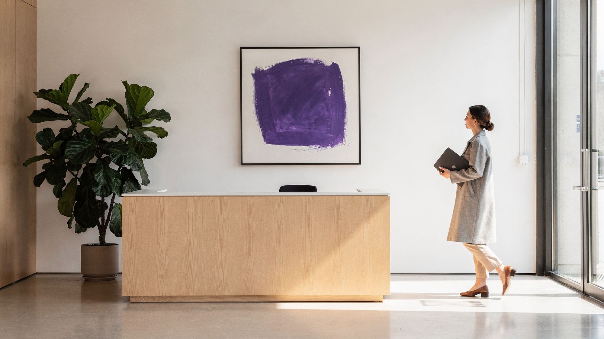 A woman carrying a laptop walks into the entrance of a small modern studio, past a light wood reception desk and a large framed violet artwork on the white wall.