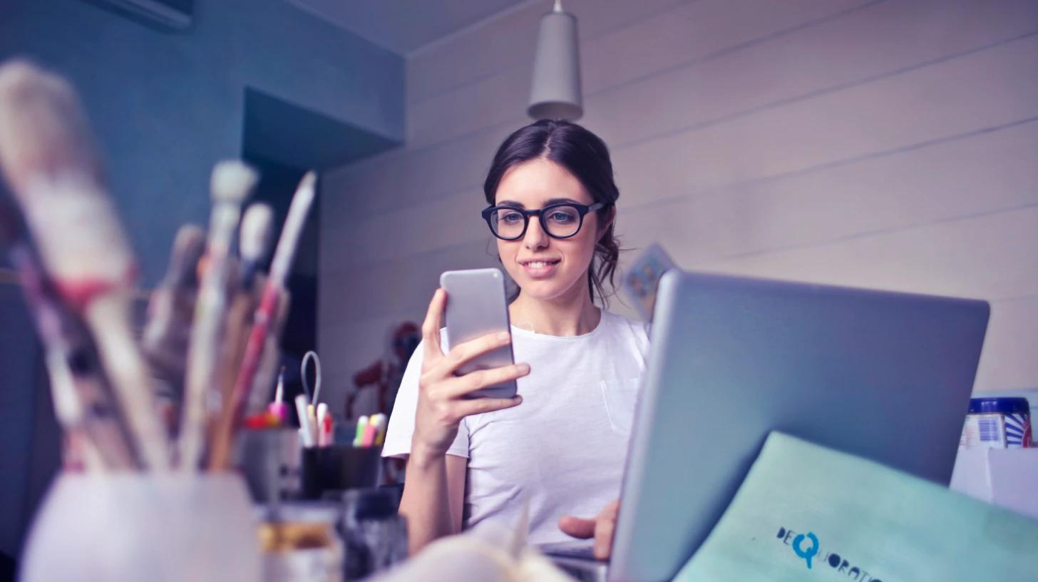 Designer wearing glasses works at laptop while reviewing content on her phone at a modern desk workspace.