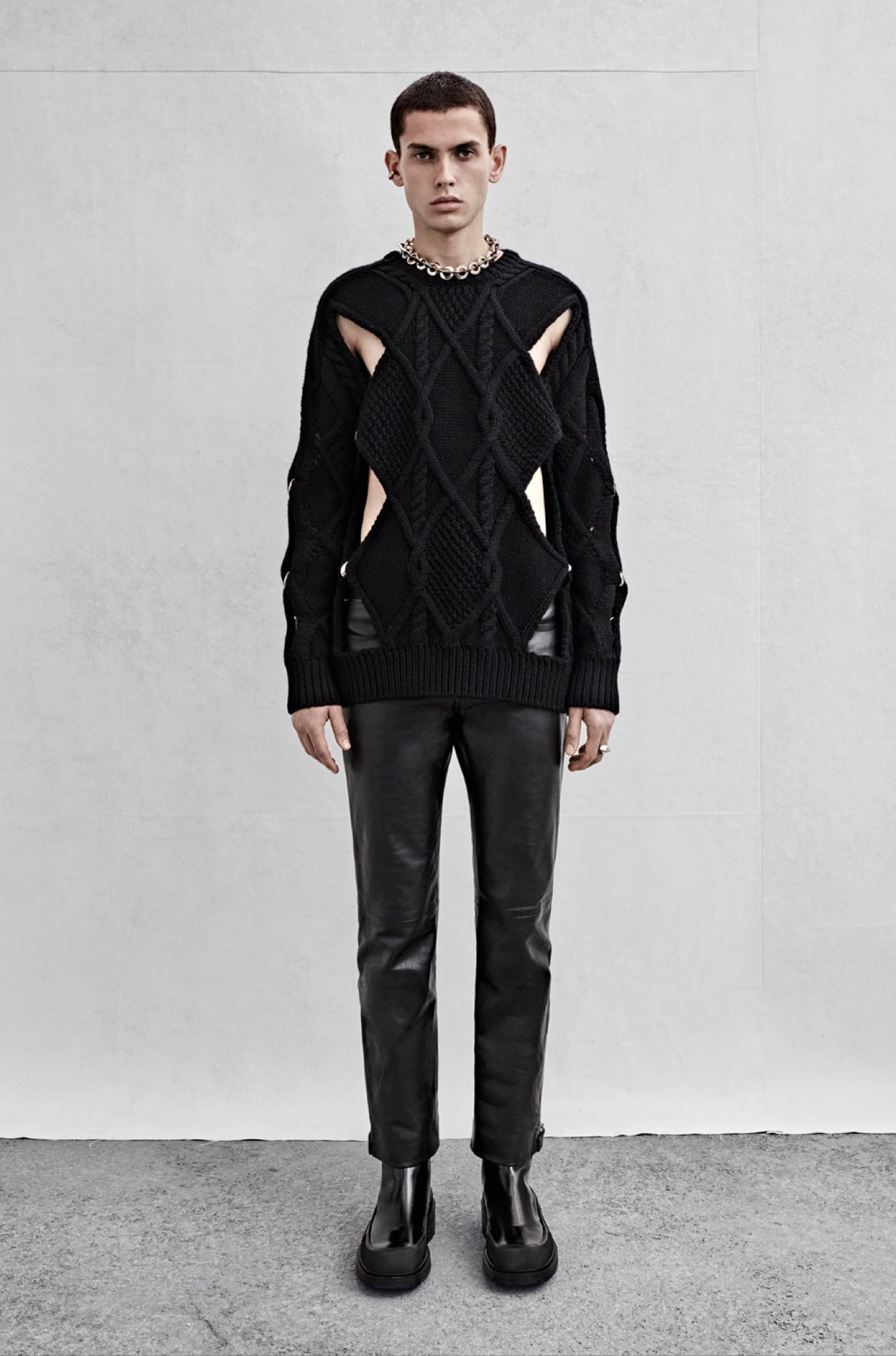 A crew neck jumper in chunky cable knit black wool with silver ring detailing and trousers in black leather with zipped hem detailing.