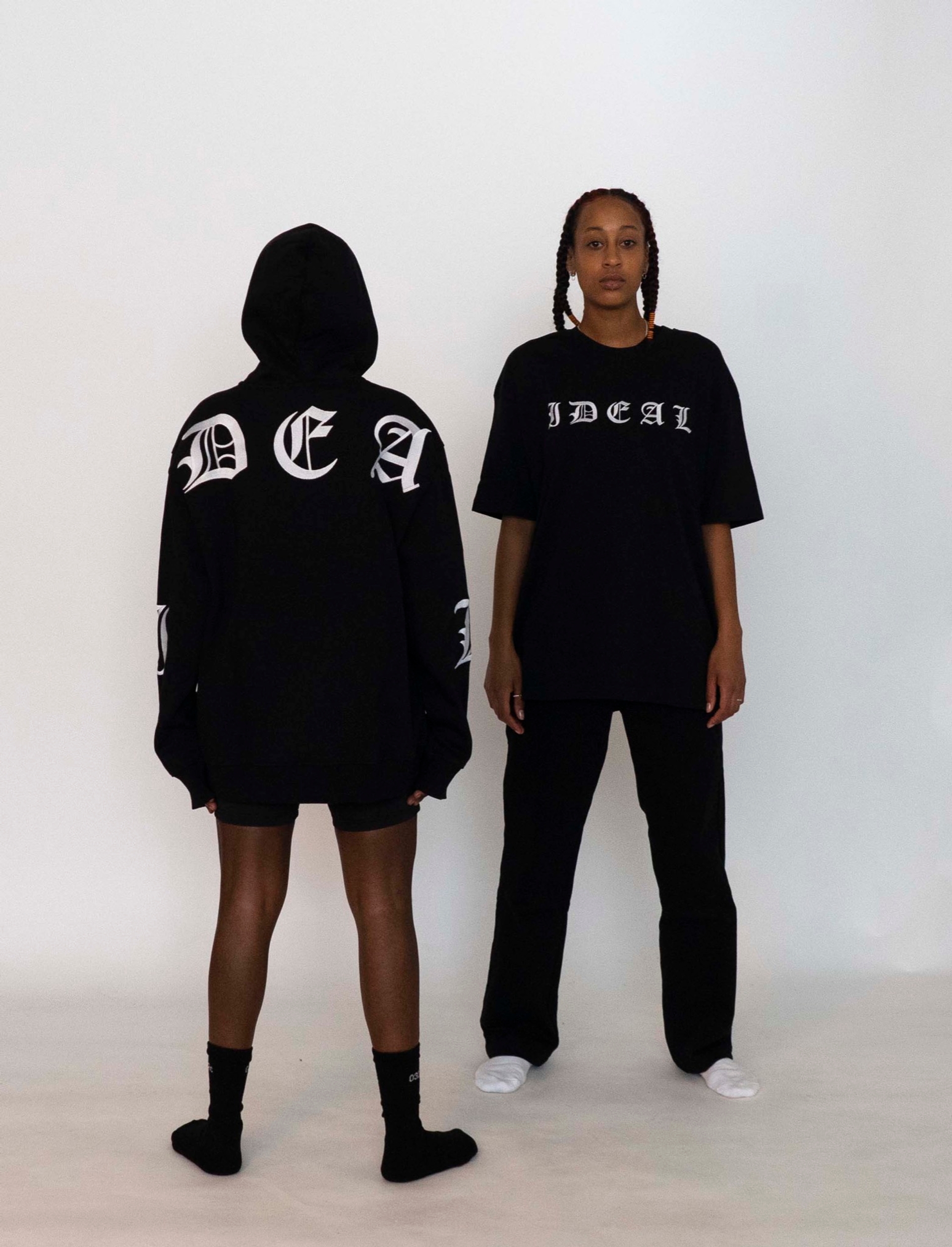 Mariama wears the 032c LoveSexDreams "Team Société"/"IDEAL" hoodie and neoprene shorts in black. Libell wears the "Ideal" t-shirt and straight leg trousers