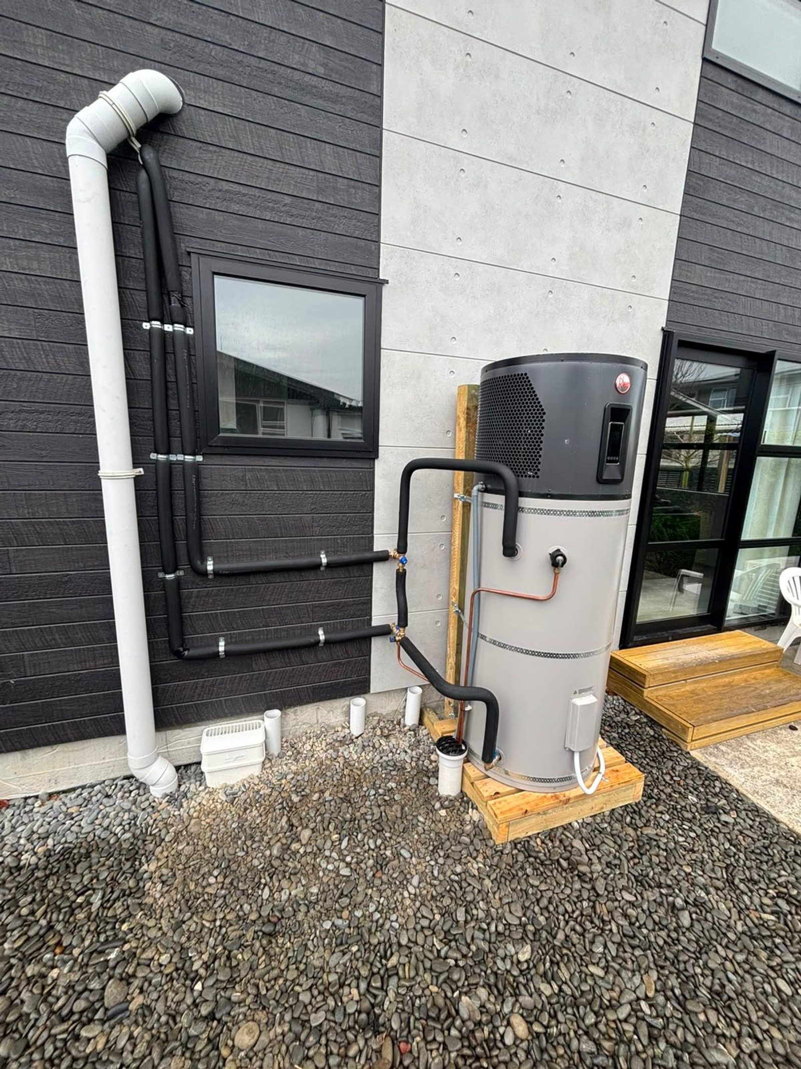 Outdoor heat pump water heater with insulated pipework beside a modern home