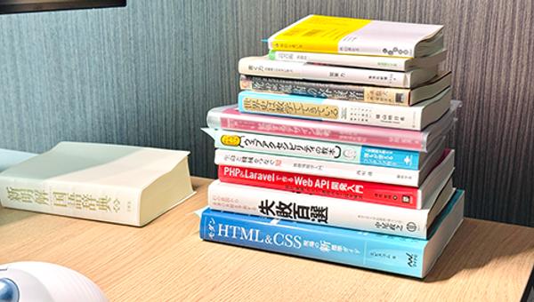 真坂の机に積まれた本の様子。技術書から教養書、小説まで幅広いジャンルの本が積まれている様子がわかる