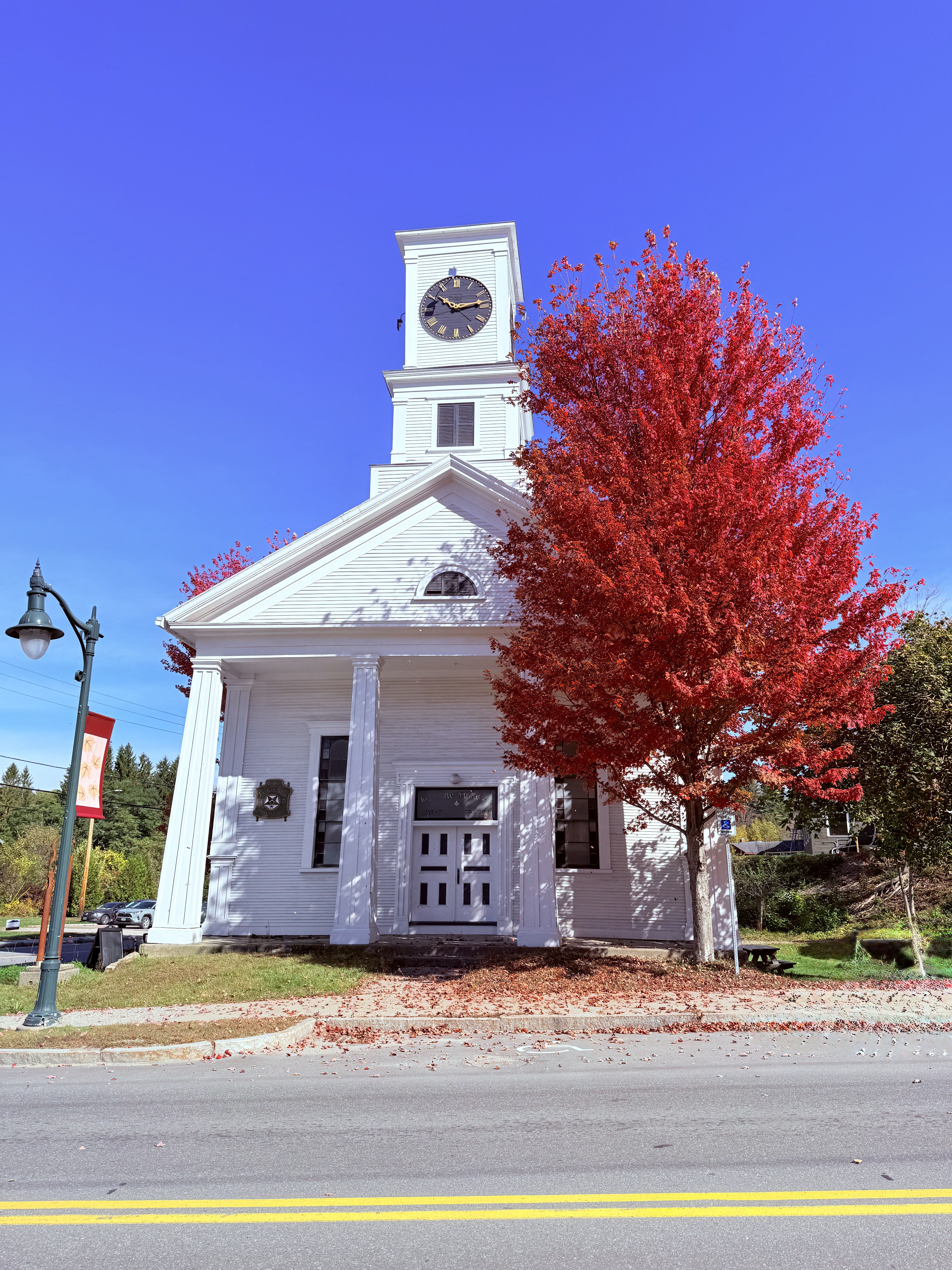 A white meeting house with a clock tower and columned front, beside a bright red maple tree under a clear blue sky.