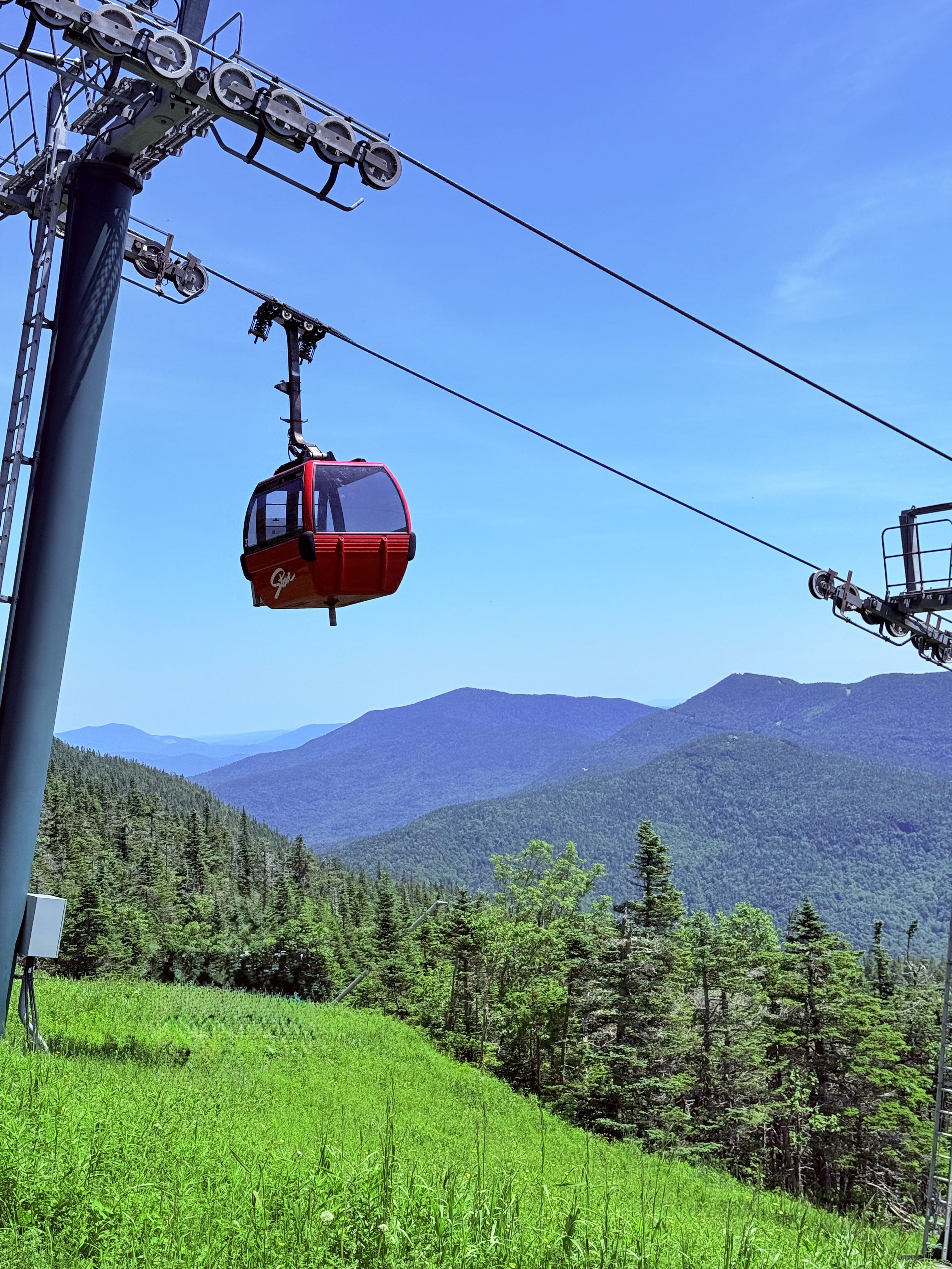 A red gondola cabin on a cable above a green summer ski slope, with forested mountain ridges and blue sky behind.