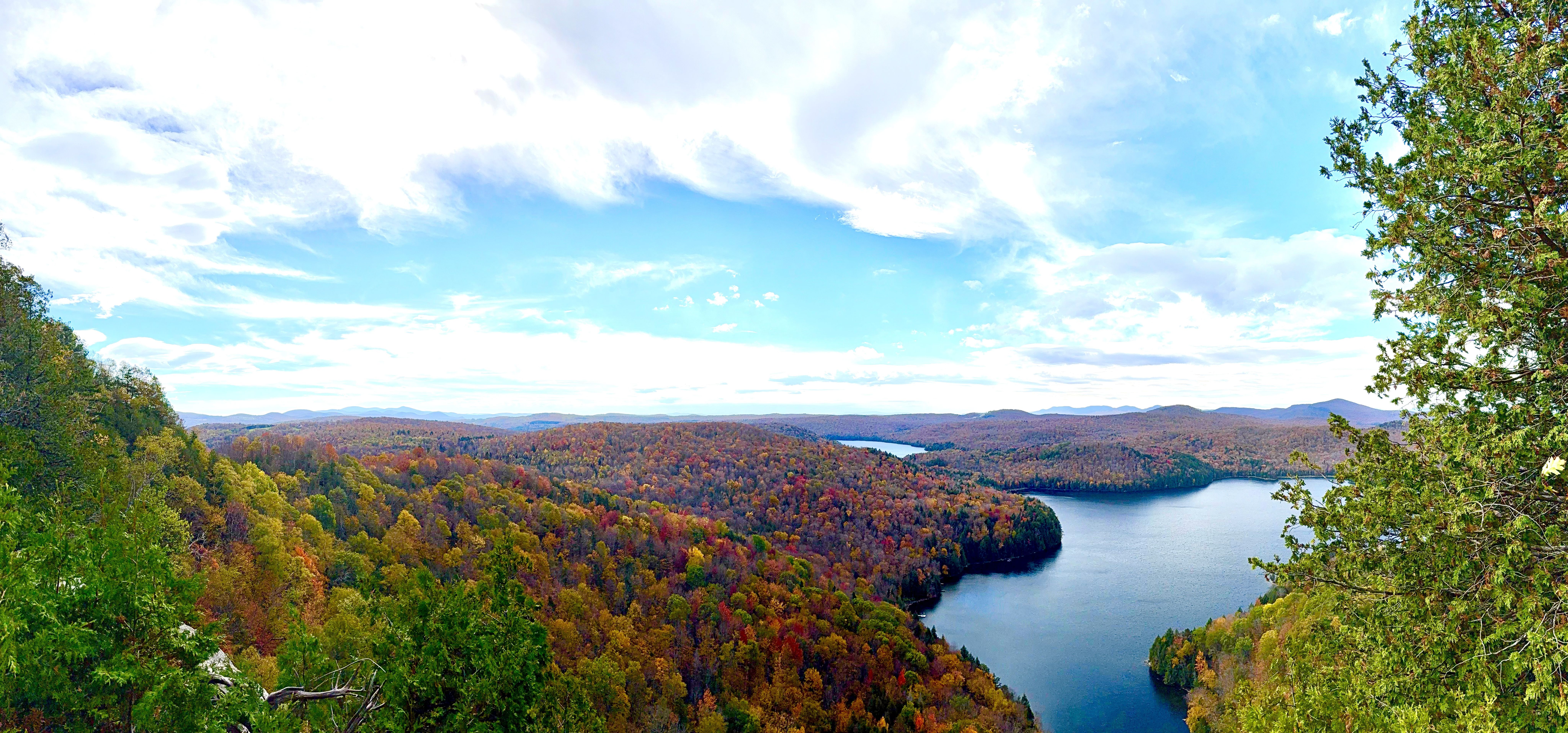 A panoramic view from a wooded overlook of a winding lake surrounded by hills in full fall color under a partly cloudy sky.