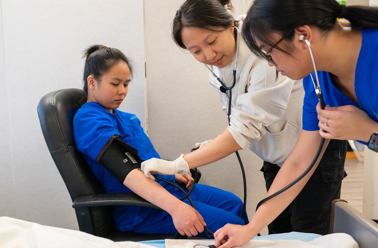 Three nursing assistant students practicing a blood pressure check at a skills lab bed