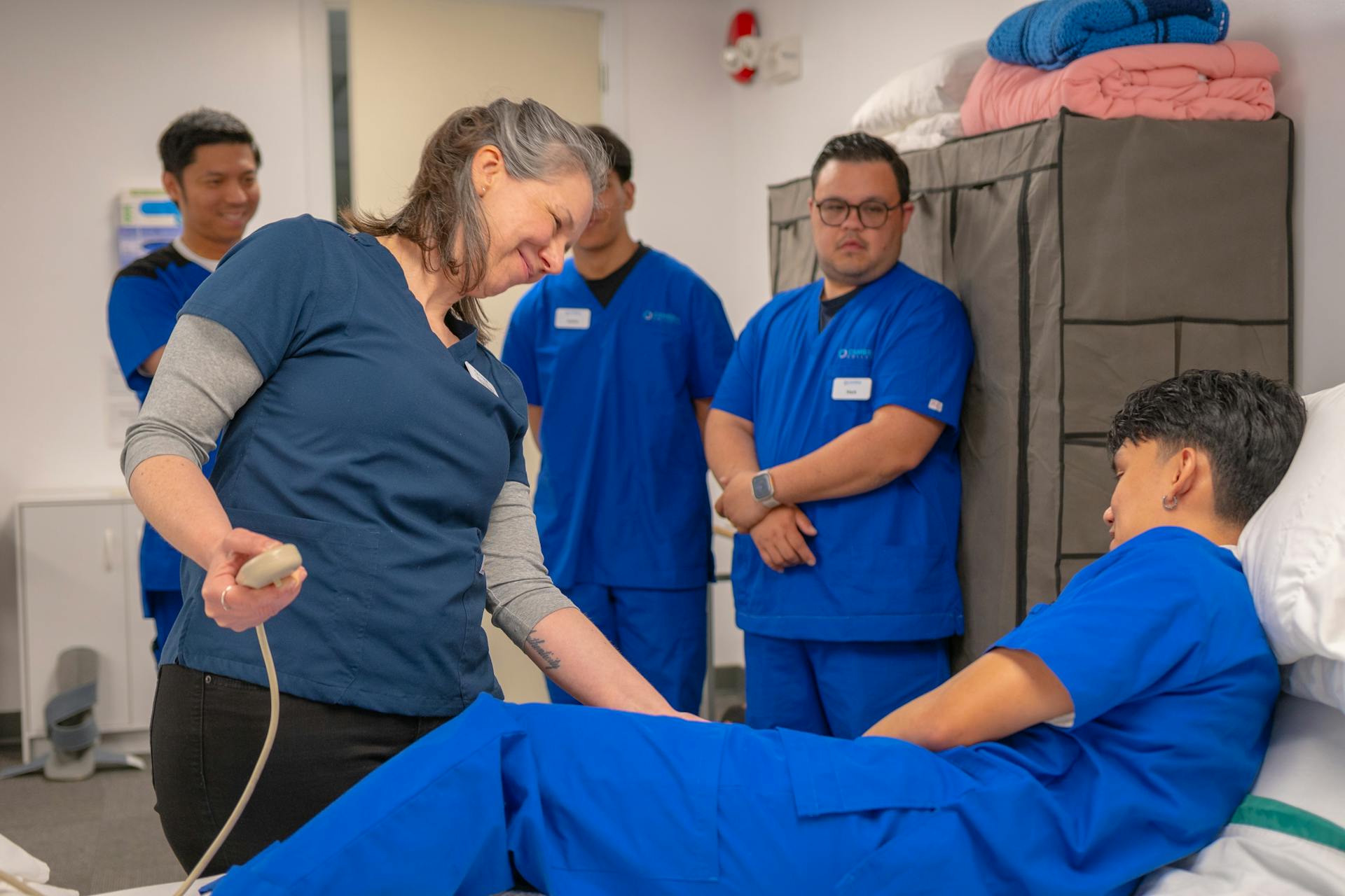 An instructor talking with a student playing the patient in a skills lab while classmates look on