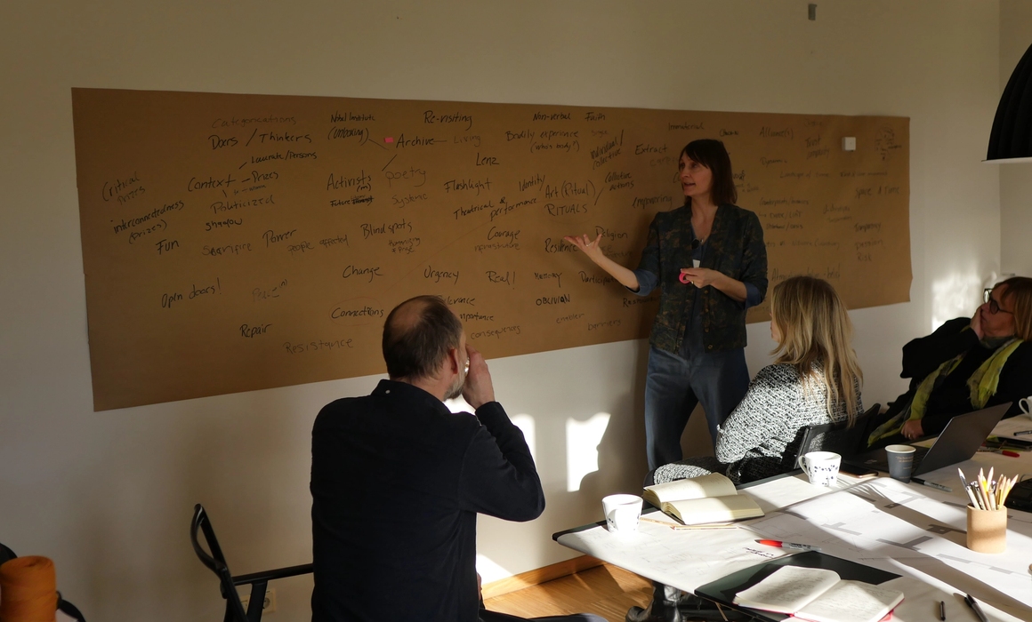 Claire speaking and showing a board with many words, people around paying attention.