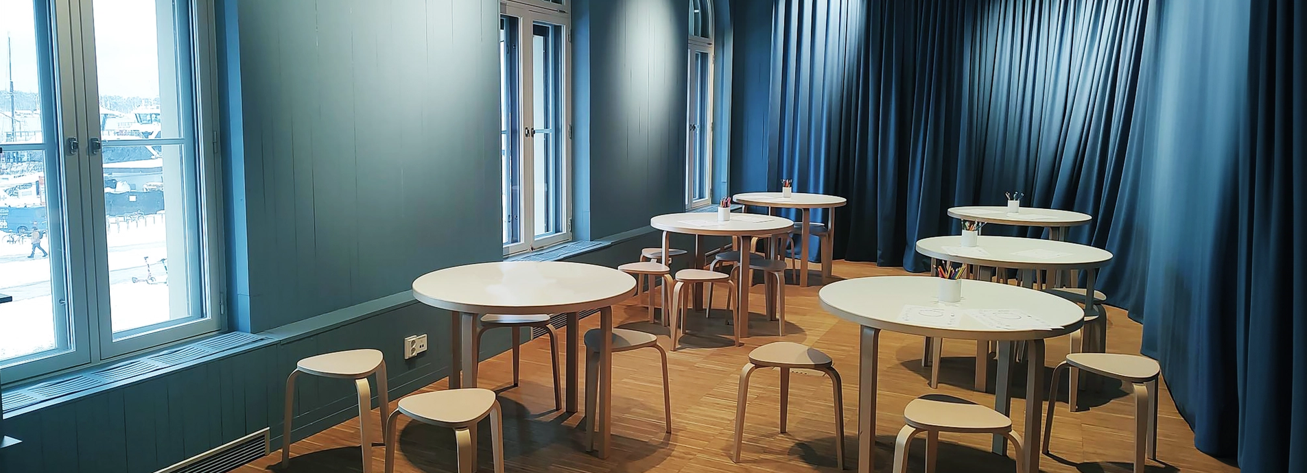 Showing the meeting room Malala. 6 round tables with backless chairs surrounding them. The room has blue walls and blue modern curtains as well as big light windows. Perfect for low formality gatherings.