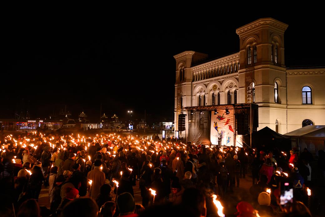 A big crowd with lit torches in front of the nobel peace center and a stage in evening time.