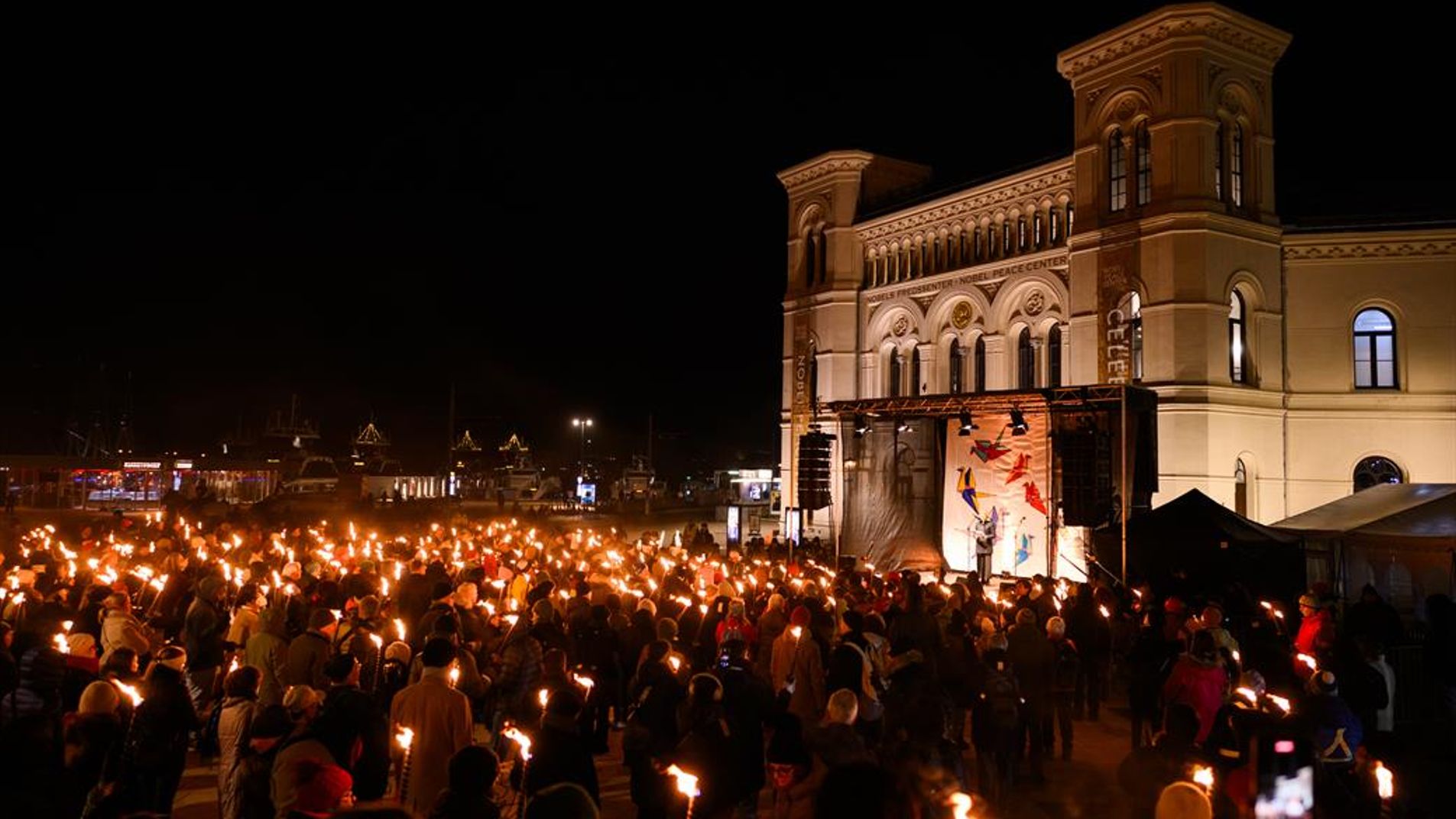 A big crowd with lit torches in front of the nobel peace center and a stage in evening time.
