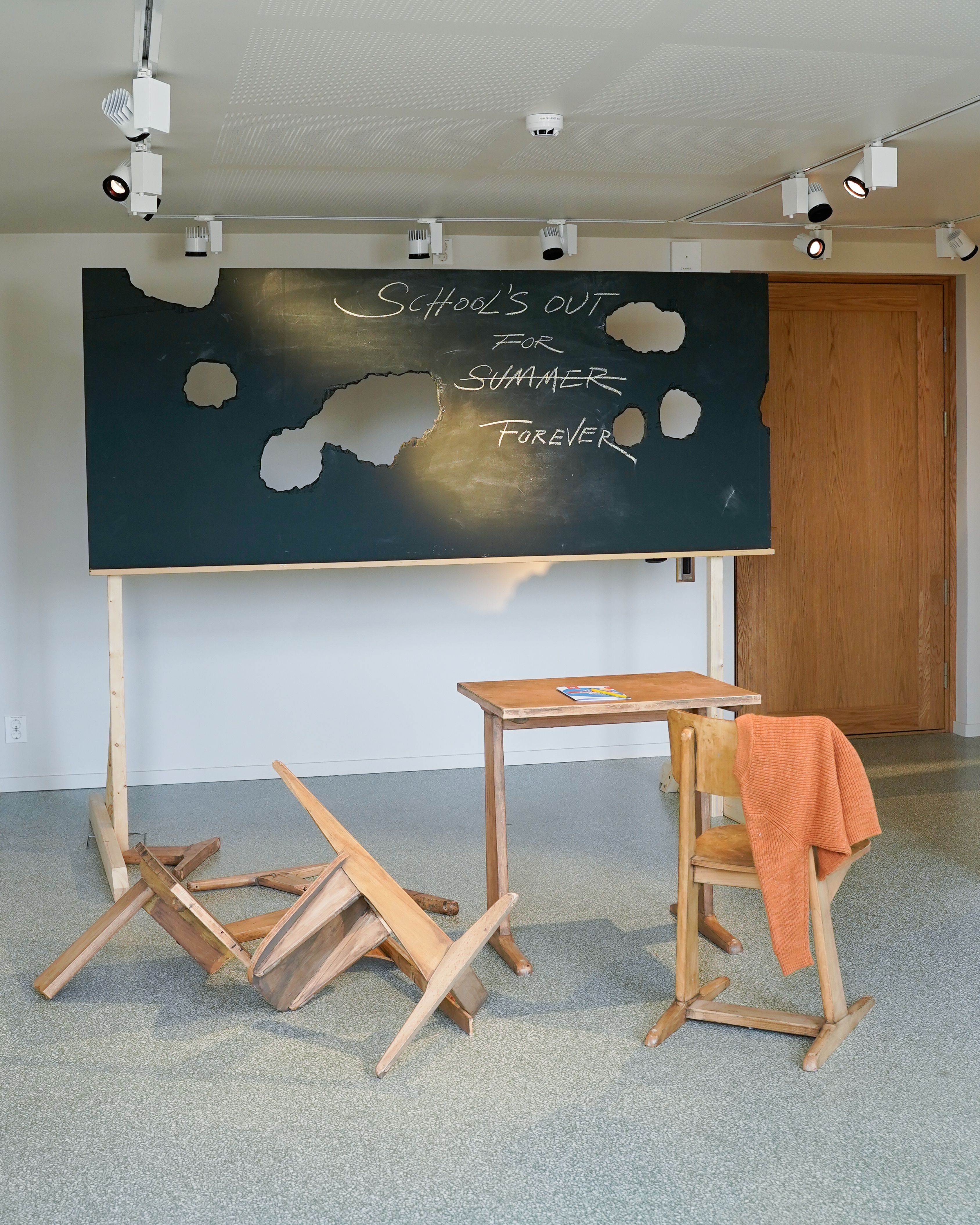 Fra utstillingen Class of No Tomorrow. En skoletavle med hull og skrift i. Ødelagte skolestoler.