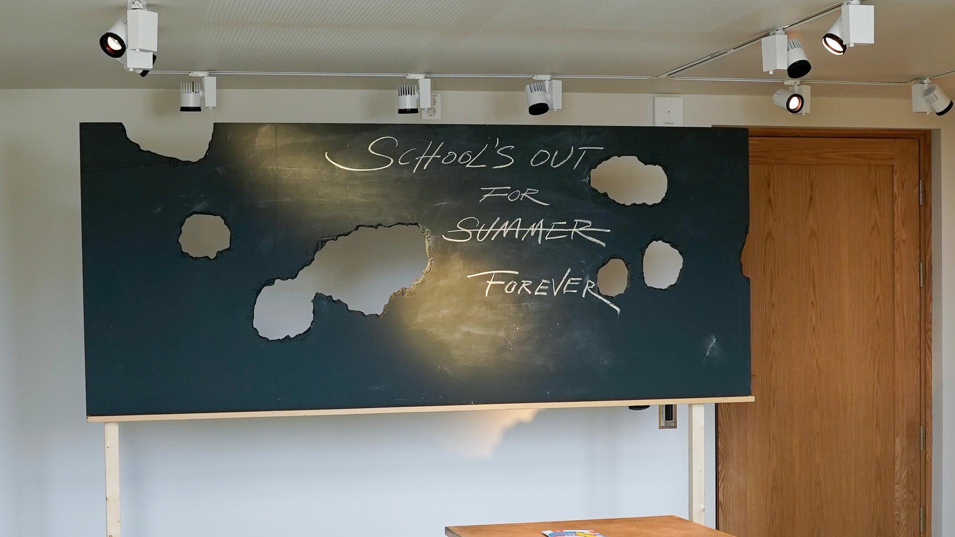 Fra utstillingen Class of No Tomorrow. En skoletavle med hull og skrift i. Ødelagte skolestoler.